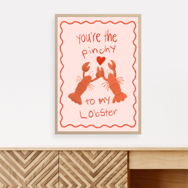 Illustration of two orange lobsters holding claws with a heart and text "You're the Pinchy to my Lobster" on a pink poster with a squiggly red outline. The framed poster is displayed on a wall with a brown chest of drawers beneath it.