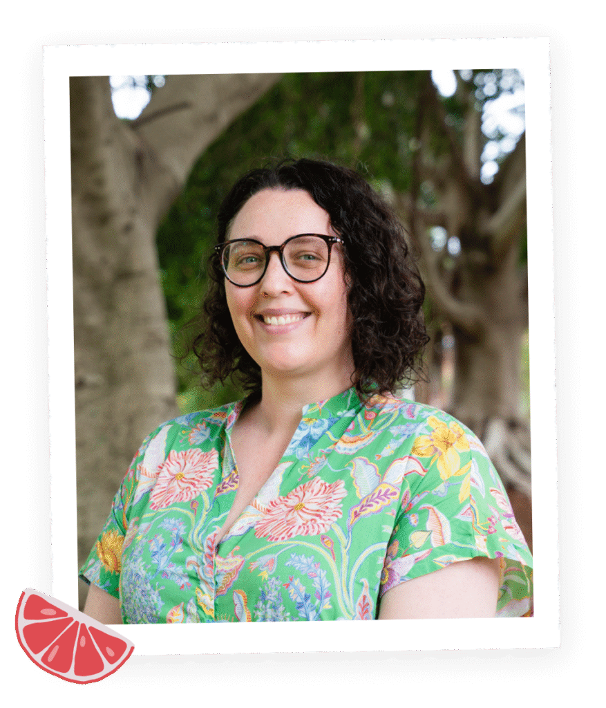 Portrait of Liv Lorkin, a talented author, illustrator, and designer. She is captured smiling at the camera, showcasing shoulder-length short brown hair and a freckled face. The background features illustrations on the wall, providing a glimpse into her creative and artistic environment.