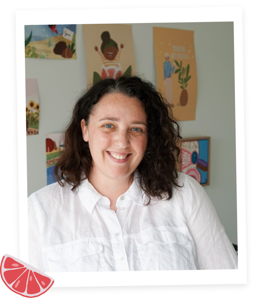 Portrait of Liv Lorkin, a talented author, illustrator, and designer. She is captured smiling at the camera, showcasing shoulder-length short brown hair and a freckled face. The background features illustrations on the wall, providing a glimpse into her creative and artistic environment.